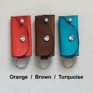 Puede incluir: Tres llaveros de cuero en naranja, marr&oacute;n y turquesa. Cada llavero tiene un anillo de metal plateado y un bot&oacute;n de metal plateado.