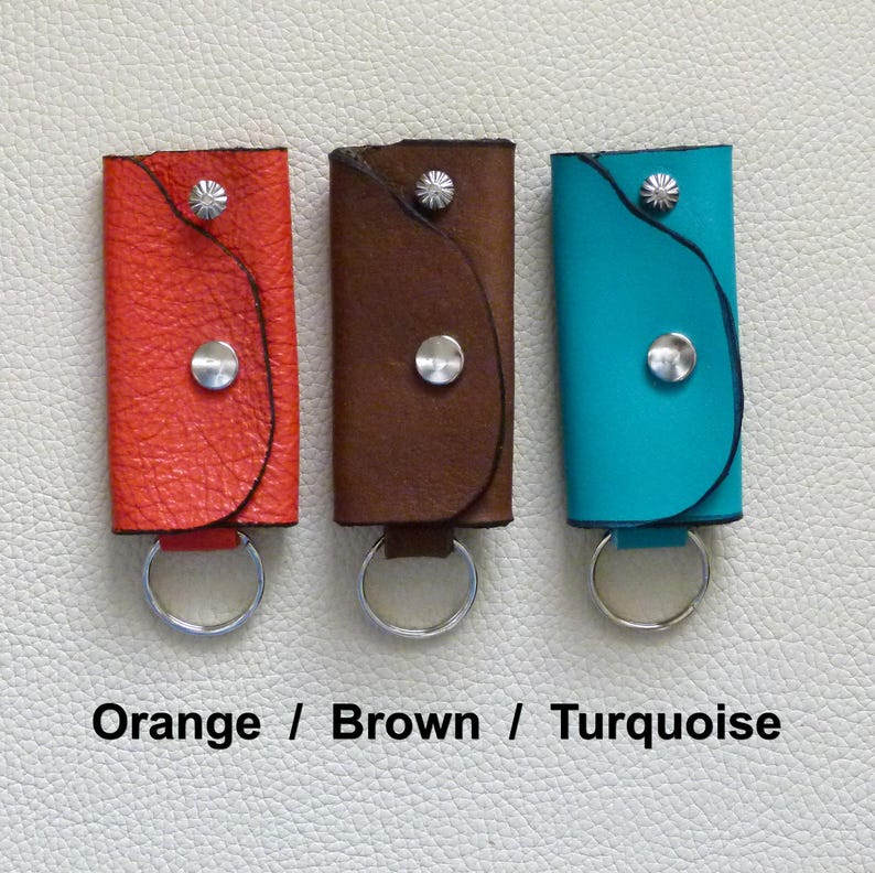 Puede incluir: Tres llaveros de cuero en naranja, marr&oacute;n y turquesa. Cada llavero tiene un anillo de metal plateado y un bot&oacute;n de metal plateado.