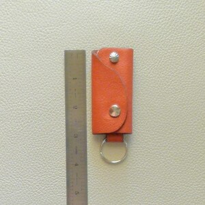Puede incluir: Porta llaves de cuero naranja con cierre de presi&oacute;n plateado y un anillo plateado. El porta llaves mide aproximadamente 5 cm de alto.