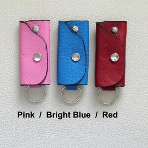 Puede incluir: Tres llaveros de cuero con anillos de metal. Los llaveros son de color rosa, azul brillante y rojo. Cada llavero tiene un bot&oacute;n plateado en la parte delantera.