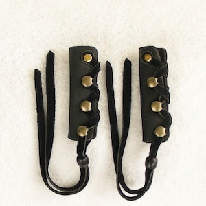 May include: Two black leather wrist cuffs with gold metal accents and black tassels. The cuffs are laced together with black cord.