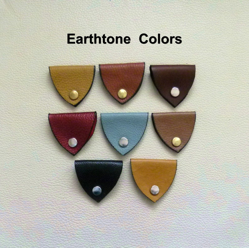May include: A set of ten leather cord keepers in various earthtone colors. The cord keepers are shaped like a triangle with rounded edges. Each cord keeper has a small metal ring in the center.