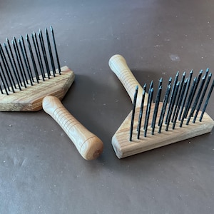May include: Three wooden wool combs with black metal tines. The combs have light-coloured wooden handles and bases. The tines are evenly spaced and appear sharp. The combs are arranged on a brown surface.