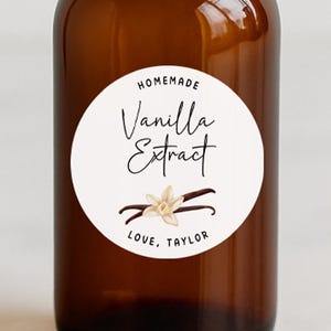 May include: A brown glass bottle with a black cap, labeled "Homemade Vanilla Extract." The white label features the text "Vanilla Extract" in elegant script, with a vanilla flower illustration. The words "Love, Taylor" are at the bottom.