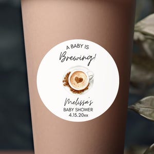 May include: A brown paper coffee cup with a white lid and a circular sticker that reads "A Baby Is Brewing!" with a coffee cup graphic. Coffee beans are scattered at the base of the cup. The background features green leaves.