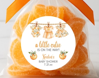 May include: A clear cellophane bag filled with orange slices, tied with an orange ribbon. A round sticker on the bag reads "a little cutie is on the way!" with baby clothes illustrations and baby shower details.