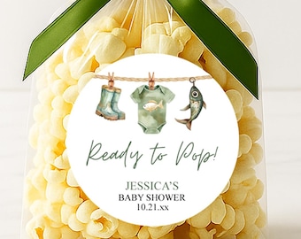 May include: A clear cellophane bag filled with popcorn, tied with a dark green ribbon. A round sticker with baby-themed illustrations and the text "Ready to Pop!" is attached to the bag. The sticker includes a baby boot, onesie, and fish.