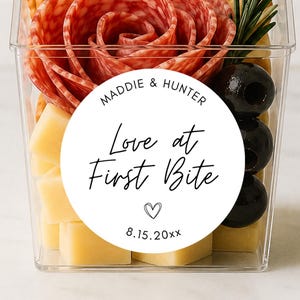 May include: A clear plastic container filled with crackers, salami folded into a rose shape, cubed yellow cheese, black olives, and a sprig of rosemary. A round white sticker features the text "Maddie & Hunter Love at First Bite" with a heart and "8.15.20xx".