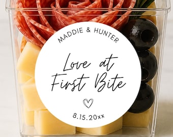 May include: A clear plastic container filled with crackers, salami folded into a rose shape, cubed yellow cheese, black olives, and a sprig of rosemary. A round white sticker features the text "Maddie & Hunter Love at First Bite" with a heart and "8.15.20xx".