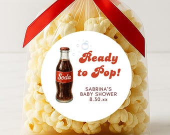Puede incluir: Una bolsa transparente llena de palomitas de maíz, atada con una cinta roja. Una pegatina redonda en la parte delantera presenta una botella de refresco y el texto "Ready to Pop!" con detalles de baby shower.