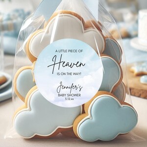 May include: A clear cellophane bag tied with a light blue ribbon, filled with cloud-shaped cookies. Each cookie has a light blue frosting and white icing. A round sticker reads "A Little Piece of Heaven is on the way!" for a baby shower.