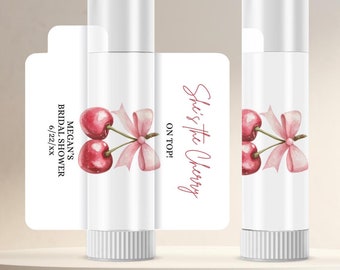 May include: White lip balm tubes with a watercolor cherry design and the text "She's the Cherry on Top!" A bridal shower theme is suggested by the text "Megan's Bridal Shower 6/22/xx". The lip balms are displayed on a light-colored platform.