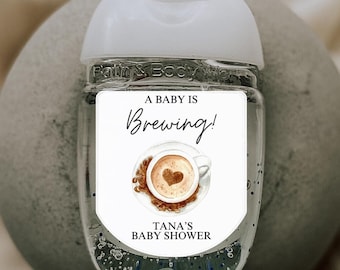 May include: A clear hand sanitizer bottle with a white cap and label. The label reads "A Baby Is Brewing!" with a coffee cup design and "Tana's Baby Shower" text. The bottle is on a gray, textured surface.