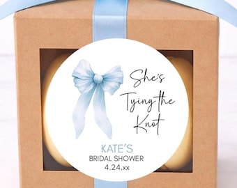 May include: A small, square, brown gift box with a light blue ribbon tied in a bow on top. A round sticker on the front reads "She's Tying the Knot" with "Kate's Bridal Shower" and the date.