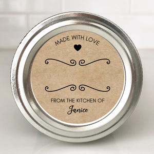 May include: A round silver metal mason jar lid featuring a circular tan paper label. The label displays the text MADE WITH LOVE, a small black heart, two decorative black scroll flourishes, and the text FROM THE KITCHEN OF Janice.