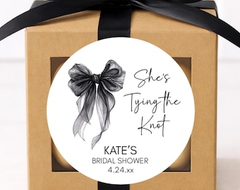 May include: A small, square, brown gift box with a black satin bow. A white circular sticker on the front reads "She's Tying the Knot" with a watercolor bow illustration and bridal shower details.