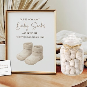 May include: A framed sign with the text "GUESS HOW MANY Baby Socks ARE IN THE JAR WHOEVER COMES CLOSEST WINS!". The sign is next to a glass jar filled with baby socks and two small cards. The theme is a baby shower game.