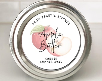 May include: A small, round, metal jar of apple butter. The label features a watercolor illustration of a pink apple and the words "Apple Butter" in cursive. Text on the label reads "From Abbey's Kitchen" and "Canned Summer 2025."