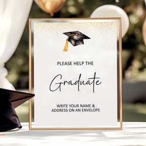 May include: A framed sign with gold glitter accents and a black graduation cap illustration, featuring the text: PLEASE HELP THE Graduate WRITE YOUR NAME & ADDRESS ON AN ENVELOPE. A black graduation cap with a gold tassel sits beside the frame.