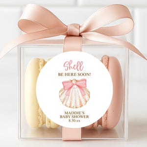 May include: A clear box tied with a pink ribbon, containing two macarons. A round sticker on the box reads "Shell be here soon!" with a seashell graphic and "Maddie's Baby Shower 8.30.xx". One macaron is yellow, the other is pink.