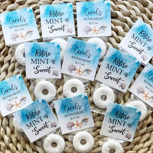 May include: Small clear bags containing white mint candies, each with a printed tag. The tags have a watercolor blue background with text that reads "Retire MINT is Sweet" and "Carol's Retirement 8.17.xx" with seashell and starfish graphics. The items are arranged on a woven surface.