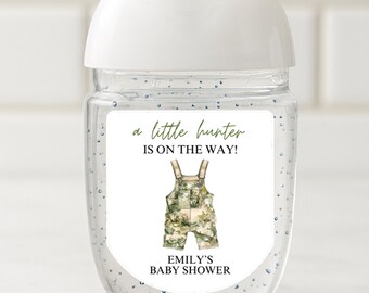 May include: A clear hand sanitizer bottle with a white cap. The label reads "a little hunter is on the way!" with an illustration of camouflage overalls. The text "EMILY'S BABY SHOWER" is below the overalls.
