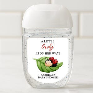 May include: Clear hand sanitizer bottle with a white top and a label. The label features the text "A LITTLE LADY IS ON HER WAY!" and a cartoon ladybug on a green leaf. The words "SABRINA'S BABY SHOWER" are also on the label.