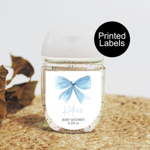 May include: A clear hand sanitizer bottle with a white cap and a label featuring a light blue watercolor bow. The label also includes the text "Katie's BABY SHOWER 4.24.xx". A black circle with the words "Printed Labels" is in the upper right.
