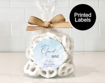 May include: A cellophane bag filled with white chocolate-covered pretzels, secured with a brown ribbon. The bag features a round label with the text "WE ARE ON Cloud NINE" and "Shelley's BABY SHOWER 3.15.25". A black circle with "Printed Labels" is also visible. The background is a white tiled wall.