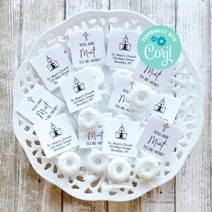 Puede incluir: Un plato blanco con pequeñas bolsas blancas que contienen mentas redondas blancas. Cada bolsa tiene una pequeña etiqueta cuadrada blanca con texto negro que dice "You are Mint to be here!" y una pequeña ilustración en blanco y negro de una iglesia. Algunas etiquetas también incluyen el texto "St. Mary's Church, 125 Main Street, Brooklyn, NY".