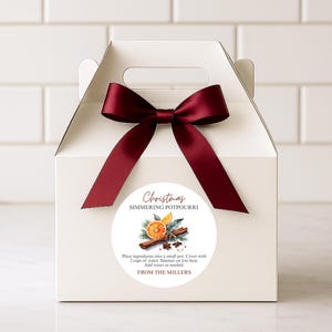 May include: A white gift box tied with a burgundy bow. The front features a round sticker with "Christmas Simmering Potpourri", instructions, and a watercolour illustration of orange slices and cinnamon sticks.