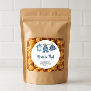 May include: A brown paper bag with a clear window filled with caramel popcorn. A round sticker on the bag reads "Ready to Pop!" with baby clothes illustrations and the text "Sabrina's Baby Shower 10.21.xx".