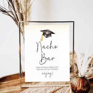 Puede incluir: Un cartel blanco con brillo dorado de fondo. El cartel dice "Nacho Bar" en escritura cursiva negra. Hay una gorra de graduación negra con una borla dorada sobre el texto. El cartel también dice "Toma tus propias patatas fritas y apila las coberturas ¡disfruta!"