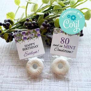 Puede incluir: Dos caramelos de menta blancos en envases de plástico transparente, con dos tarjetas pequeñas. Una tarjeta dice "Happy Birthday Evelyn!" y la otra dice "80 and in MINT Condition!" Acentos florales morados y verdes en la parte superior de las tarjetas.
