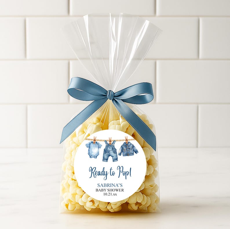 May include: A clear cellophane bag filled with popcorn, tied with a blue satin ribbon. A round sticker features watercolor illustrations of baby clothes hanging on a clothesline, with the text "Ready to Pop!" and baby shower details.