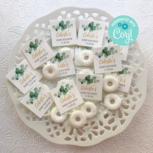 May include: A white plate with a decorative lattice pattern holds small clear bags filled with white round mints and green and gold square cards with the text "Celeste's Baby Shower 11.29.20".