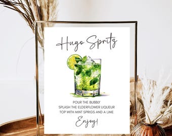 May include: A framed sign with the text "Hugo Spritz" and a watercolor illustration of a cocktail. The recipe includes elderflower liqueur, mint sprigs, and lime. The sign is in a gold frame, with decorative elements in the background.