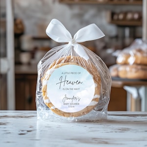 May include: A clear cellophane bag tied with a white ribbon, containing two round cookies. Each cookie has white icing and a circular label with the text "A LITTLE PIECE OF Heaven IS ON THE WAY!" and "Jennifer's BABY SHOWER 5.12.xx".