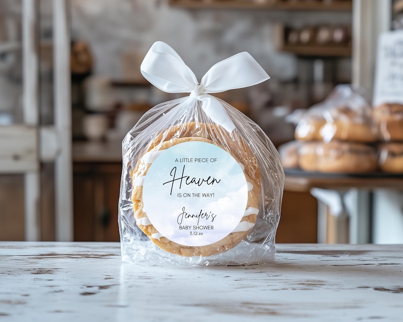 May include: A clear cellophane bag tied with a white ribbon, containing two round cookies. Each cookie has white icing and a circular label with the text "A LITTLE PIECE OF Heaven IS ON THE WAY!" and "Jennifer's BABY SHOWER 5.12.xx".