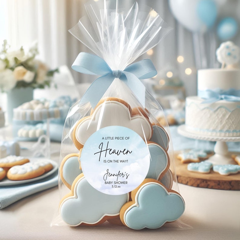 May include: A clear cellophane bag tied with a light blue ribbon, filled with cloud-shaped cookies. Each cookie has a light blue frosting and white icing. A round sticker reads "A Little Piece of Heaven is on the way!" for a baby shower.
