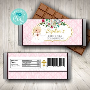 Puede incluir: Un envoltorio de barra de chocolate rosa y blanco con un diseño floral y un marco dorado. El envoltorio presenta a una niña de dibujos animados con un vestido blanco y un collar con una cruz. El texto "Sophia's First Holy Communion" está impreso en letras doradas.