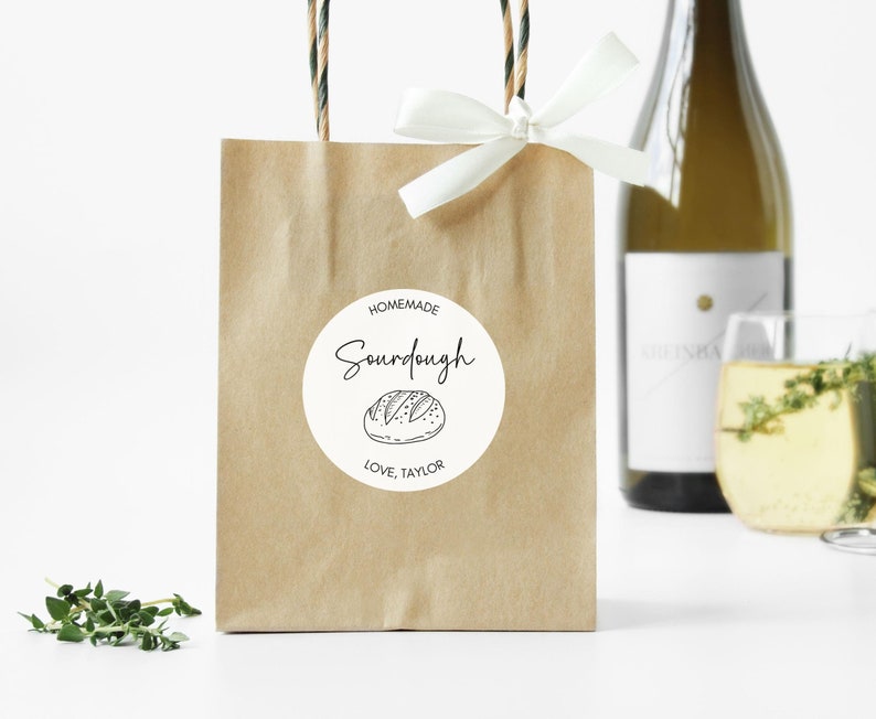 May include: A brown paper gift bag with a white bow and a round sticker that says "Homemade Sourdough" with a bread illustration. The bag has brown and green twisted paper handles. A bottle of wine and a glass of wine are in the background.
