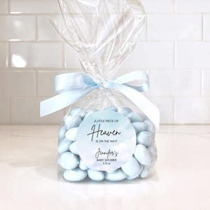 May include: A clear cellophane bag tied with a light blue ribbon, filled with light blue candies. A round sticker on the bag reads "A little piece of Heaven is on the way!" with a baby shower announcement.