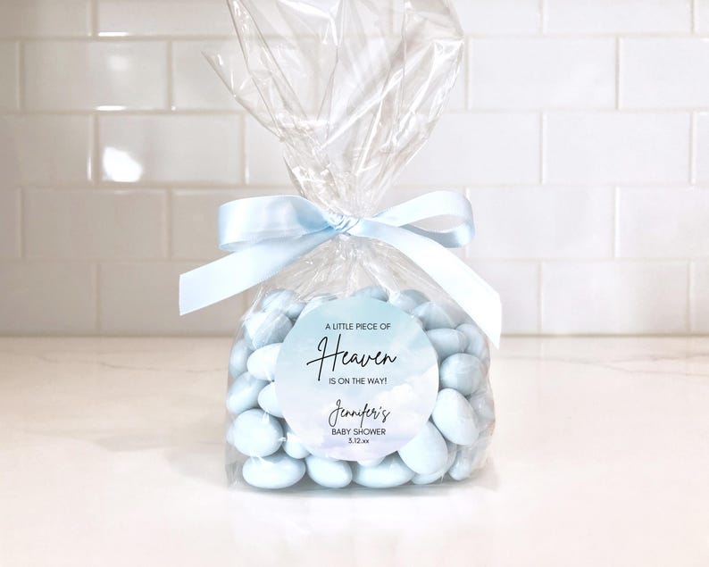 May include: A clear cellophane bag tied with a light blue ribbon, filled with light blue candies. A round sticker on the bag reads "A little piece of Heaven is on the way!" with a baby shower announcement.