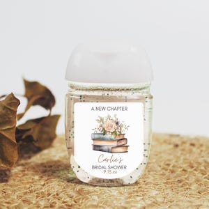 May include: Clear hand sanitizer bottle with a white cap and a label that reads "A New Chapter." The label features a floral design above a stack of books and the text "Carlie's Bridal Shower 9.15.xx."