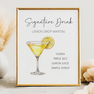 Puede incluir: Impresión enmarcada con borde dorado que muestra una ilustración de acuarela de un Lemon Drop Martini. El texto "Signature Drink" y "Lemon Drop Martini" están en la parte superior. Ingredientes: vodka, triple sec, zumo de limón y jarabe de azúcar.
