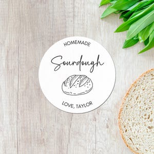 May include: A white, round label with the words "Homemade Sourdough" in black script, and a line drawing of a loaf of bread. The text "Love, Taylor" is at the bottom. A slice of bread and green leaves are in the background.