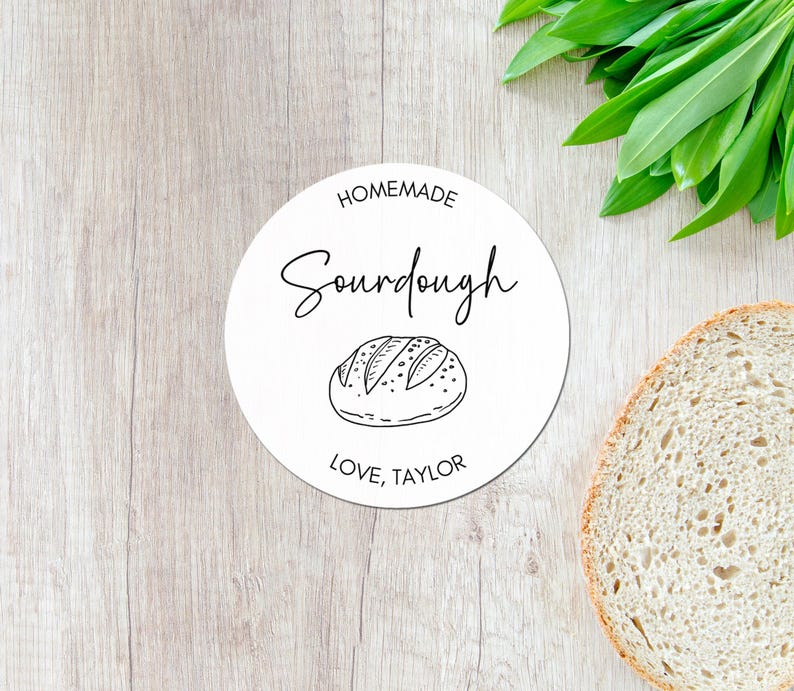 May include: A white, round label with the words "Homemade Sourdough" in black script, and a line drawing of a loaf of bread. The text "Love, Taylor" is at the bottom. A slice of bread and green leaves are in the background.