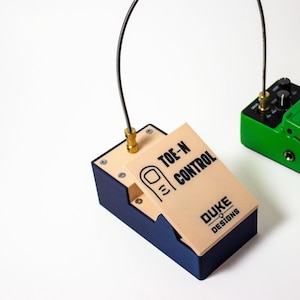 Puede incluir: Configuración de pedales de guitarra personalizados. Un pedal "Toe-N Control" azul y beige con el logo "Duke Designs" está conectado por cables a un pedal "Vintage Tube Overdrive" verde. El conjunto está sobre un fondo blanco.