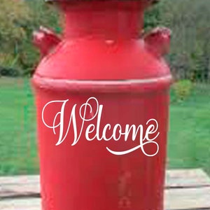 May include: Red metal milk can with a white "Welcome" sign on the front. The can is sitting on a wooden surface.